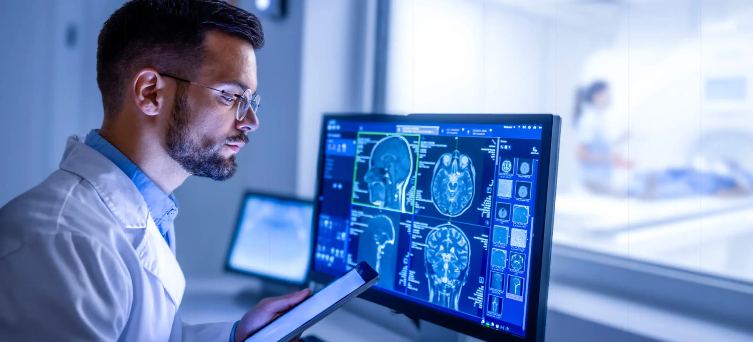 A doctor looks at a tablet while an external monitor shows a brain scan.